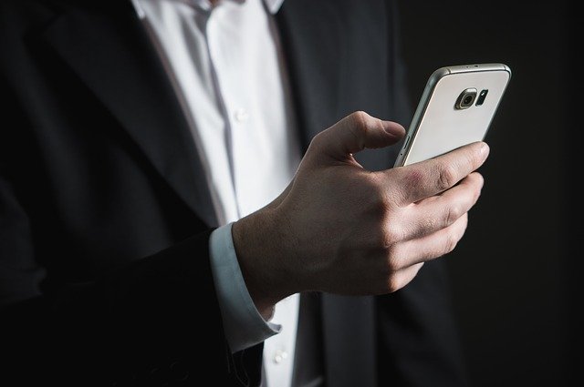 Mężczyzna w elegenckim garniturze trzyma w dłoni smartfon i kciukiem wybiera funkcję na ekranie