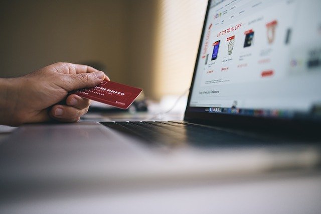 Otwarty laptop. W tle widać dłoń mężczyzny trzymającego kartę płatniczą