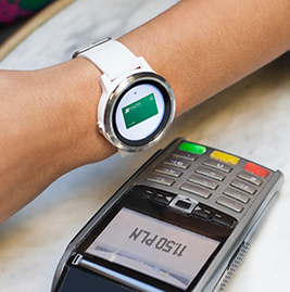 Baner reklamowy. Po lewej stronie dłoń z zegarkiem elektronicznym na nadgarstku oraz terminal płatniczy na białym tle. Na środku hasło: Czas... start, zapłacone! Błyskawiczne płatności zegarkiem. Na dole logo: Garmin Pay