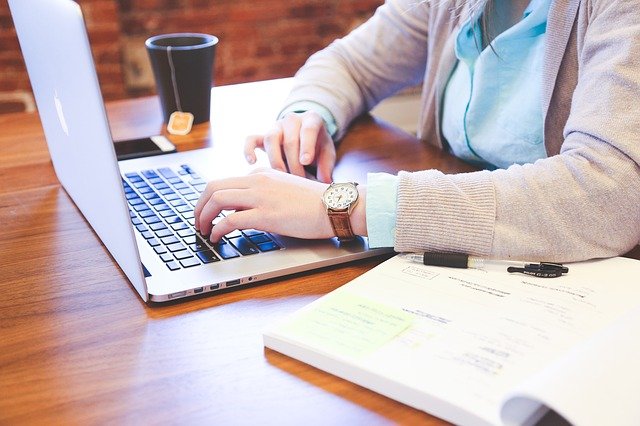 Kobieta siedzi przy stole i pracuje na laptopie. Obok niej leży otwarty notes i stoi kubek z kawą