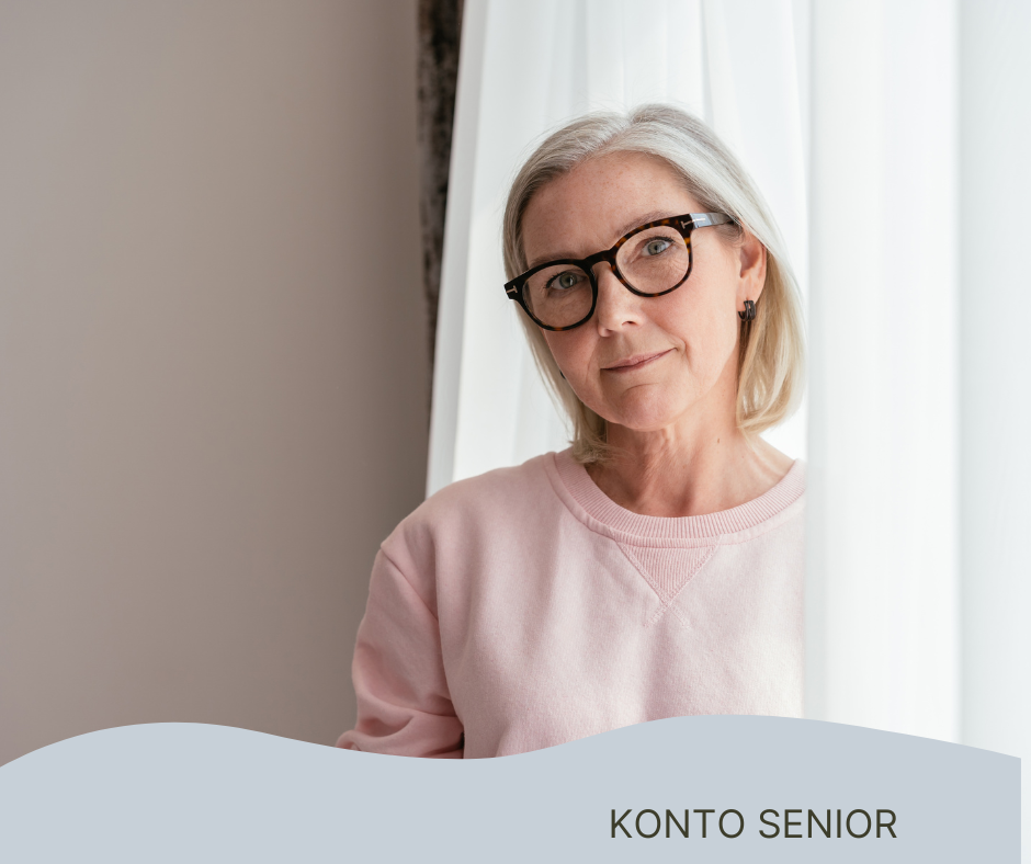 Kobieta w okularach sfotografowana na tle firanki i szarej ściany. Patrzy w obiektyw aparatu 