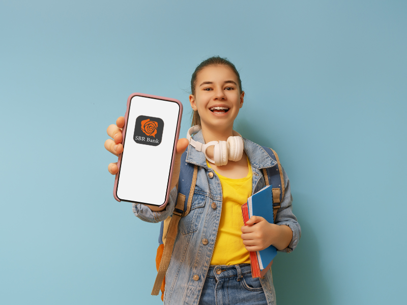 Mała dziewczynka ze szkolnym plecakiem na plecach i książkami w ręku wyciąga w stronę obiektywu aparatu smartfon. Na ekranie smartfonu widać logo SBR Bank