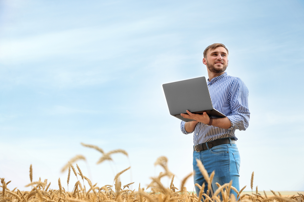 Mężczyzna stoi na polu i trzyma laptopa. Rozgląda się i nad czymś zastanawia