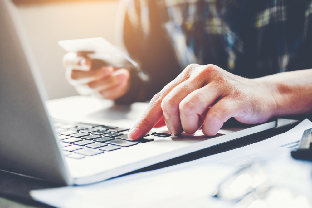 Otwarty laptop i dłonie mężczyzny pracującego na komputerze. W prawej ręce mężczyzna trzyma kartę płatniczą